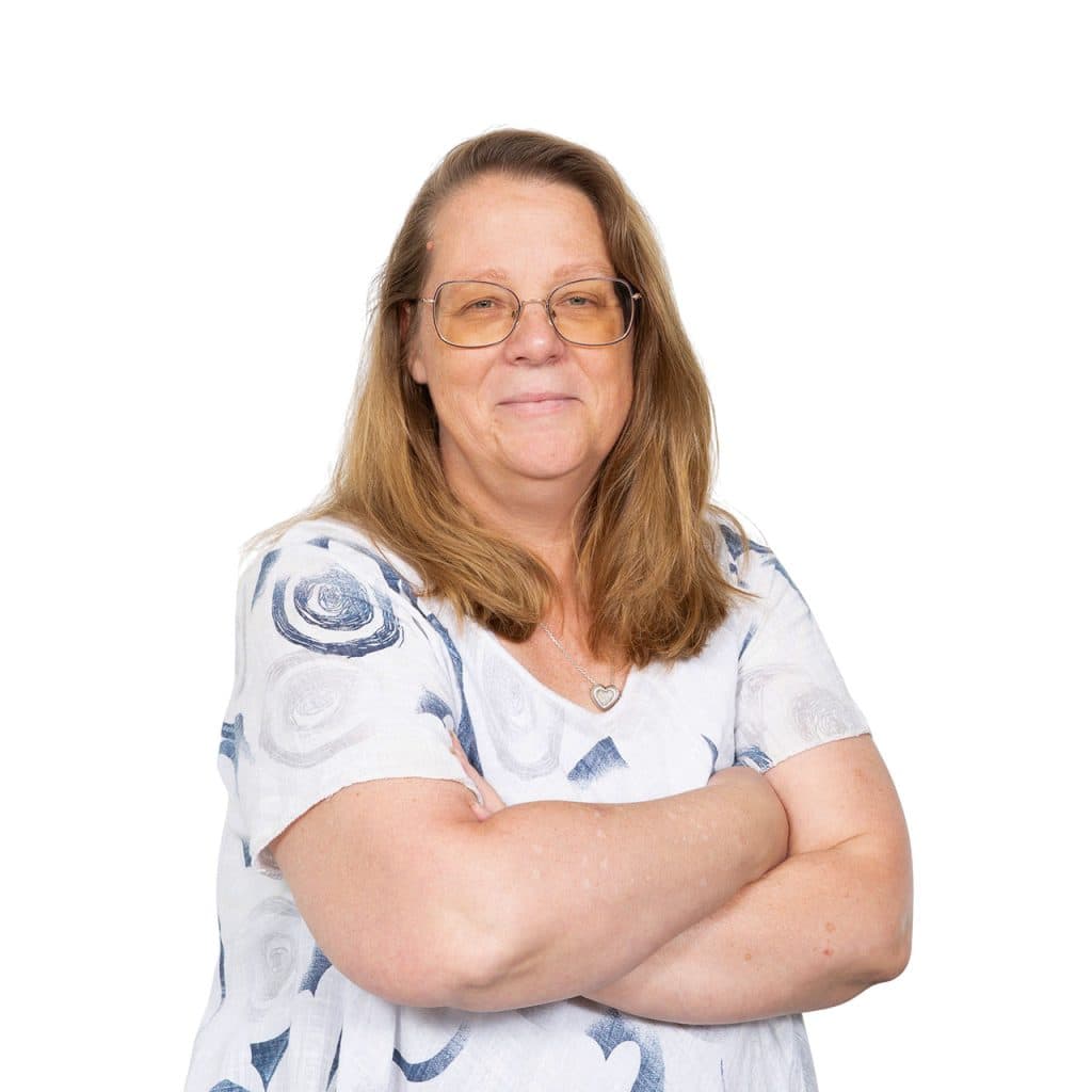 Zorgprofessional met lang blond haar en bril, gekleed in een witte blouse met blauwe patronen, met armen over elkaar, poseert zelfverzekerd tegen een witte achtergrond.