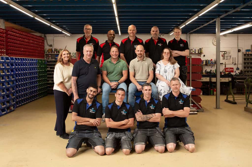 Teamfoto van Sanigas medewerkers in werkomgeving met gereedschap en opslagrekken.